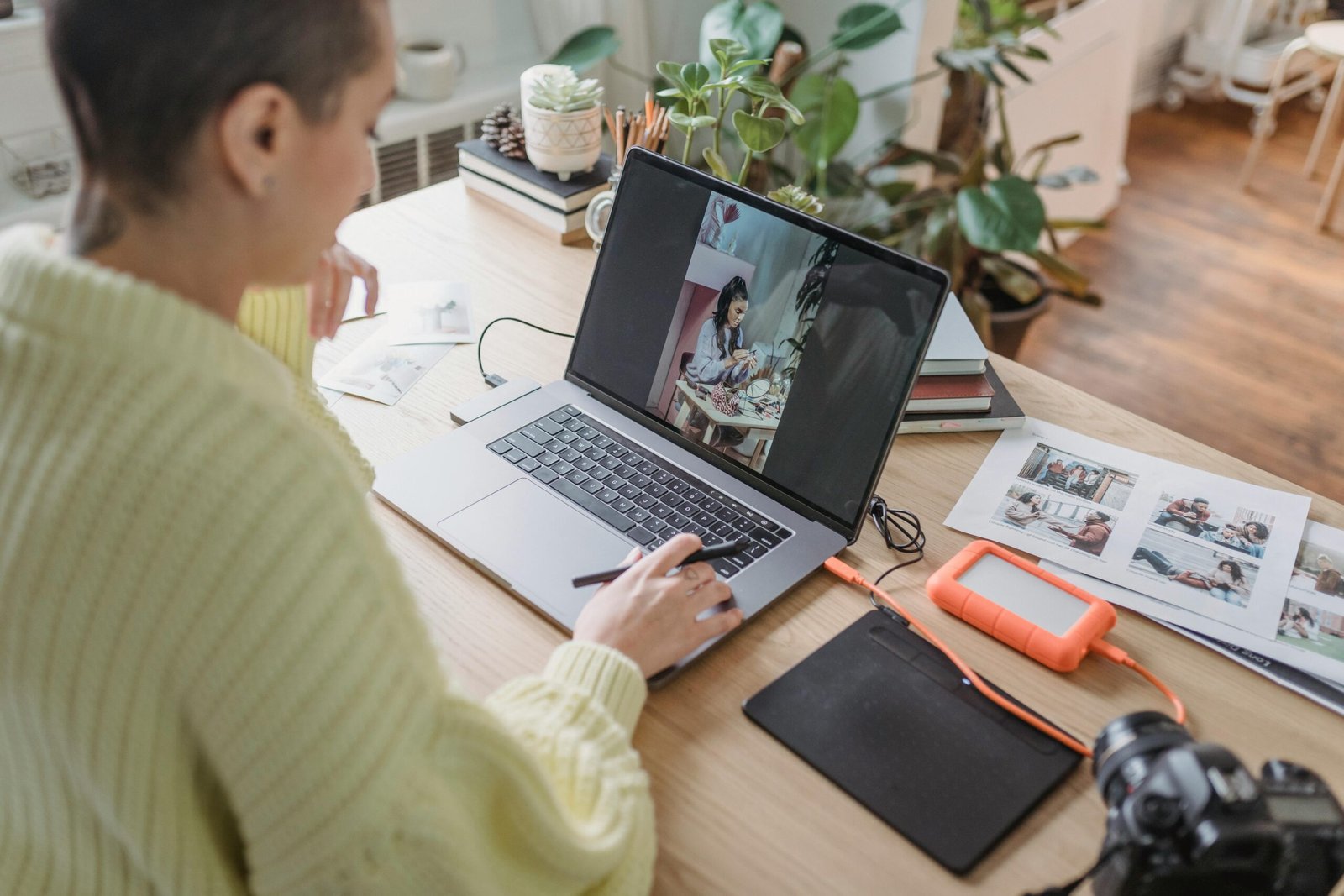 A woman edits photos on her laptop in a cozy home office, surrounded by plants and photography equipment.